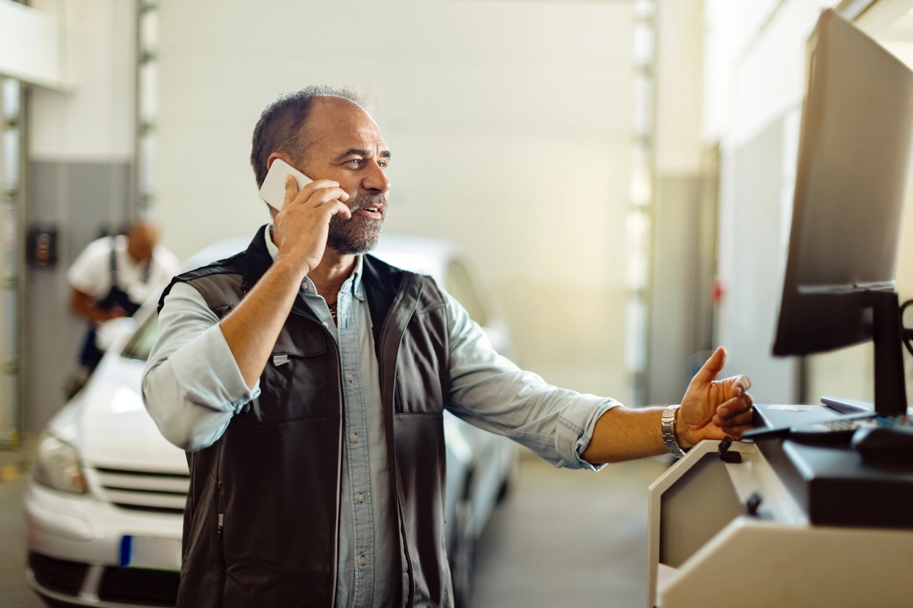 meccanico-sorridente-telefono-computer-officina-auto Meccanico sorridente che parla al telefono e utilizza un computer in un'officina automobilistica.